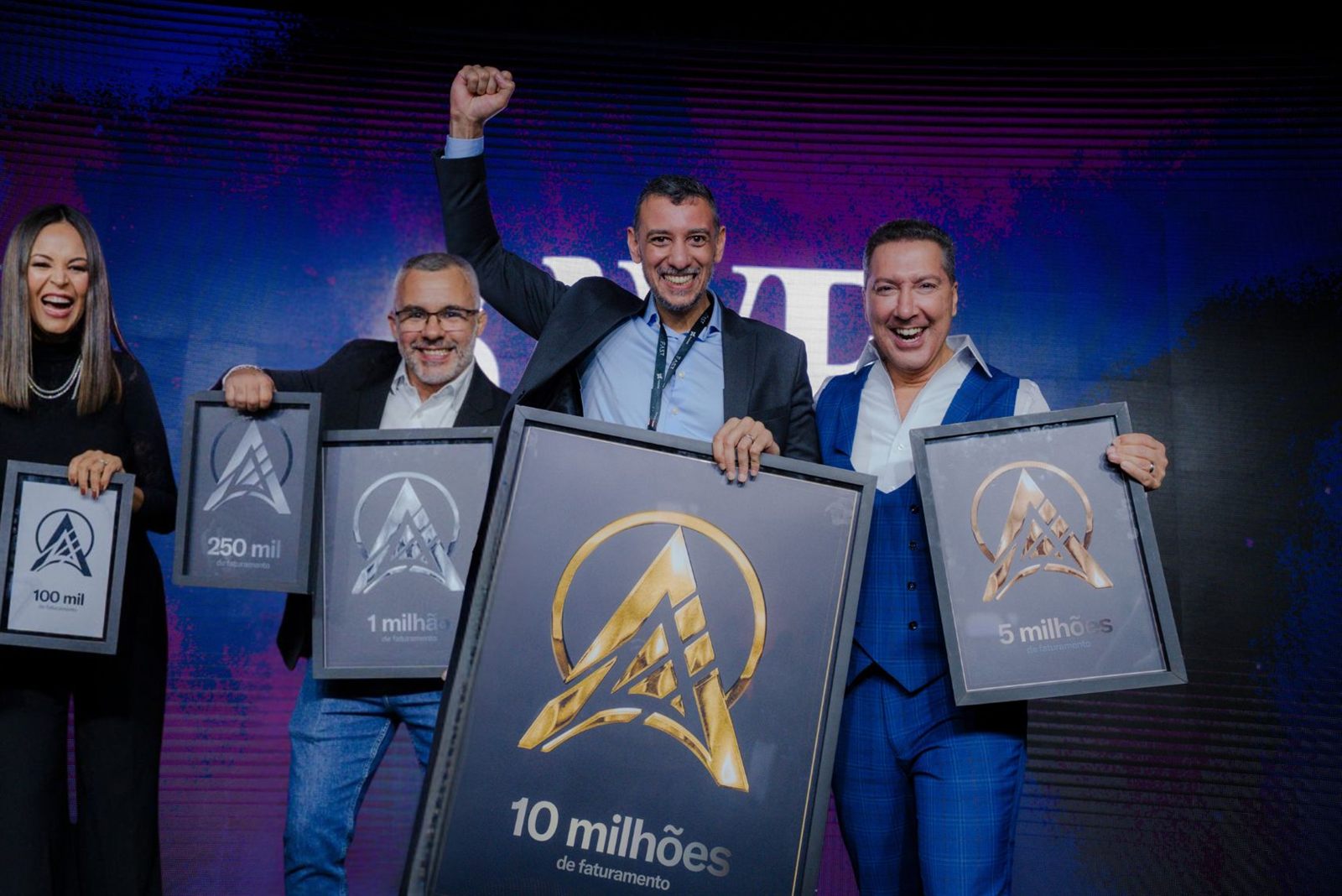 Marcos Araújo no palco do prêmio F.A.S.T., punho erguido, segurando a placa de 10 milhões de faturamento ao lado de outros premiados